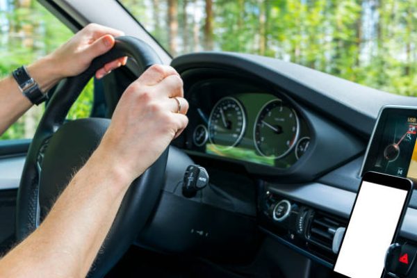 Male hands holding car steering wheel. Hands on steering wheel of a car driving near the lake. Man driving a car inside cabin. Smartphone in holder with  isolated white empty blank screen. Copy space